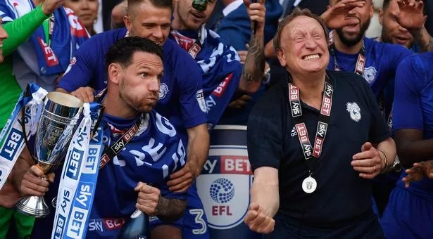A group of happy football players celebrating with a trophy, wearing blue jerseys and scarves, in a joyful team atmosphere.