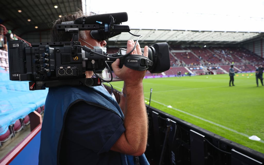 A cameraman holding a professional video camera at a sports stadium, focused on the field with empty spectator seats in the background.