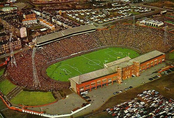 Aerial view of a crowded football stadium filled with spectators, with green pitch marked for play, surrounded by buildings and parked cars.