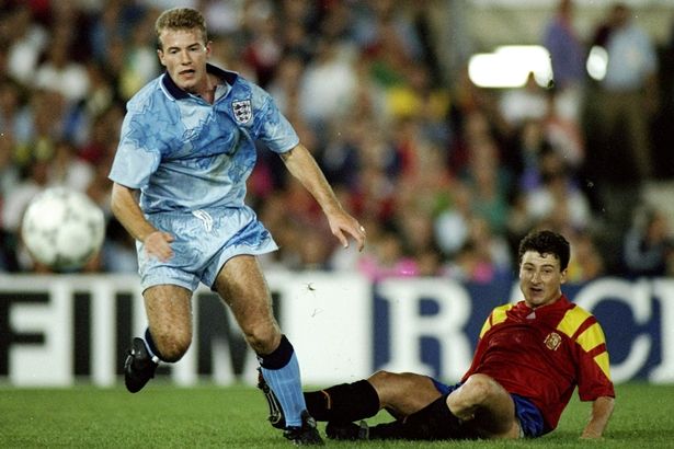 A soccer player in a light blue jersey running past a fallen opponent in a red and yellow jersey during a match.