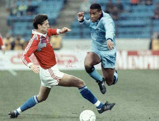 A soccer match scene featuring two players: one in a red jersey and the other in a light blue jersey, as they compete for the ball on a grassy field.